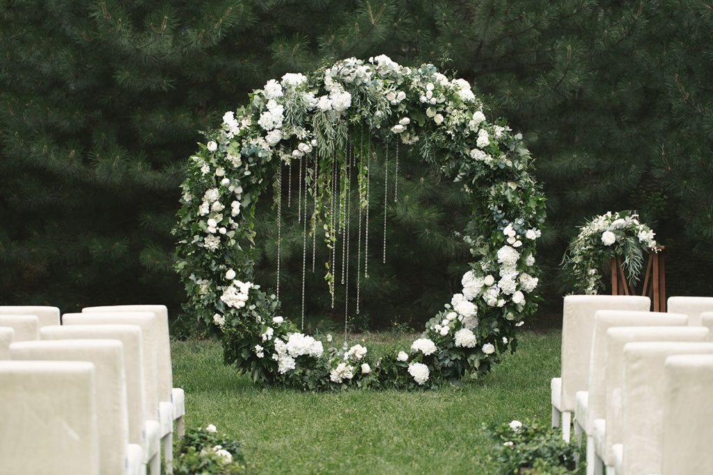 A wedding ceremony site on a grassy site, is all set with white chairs, and a larger-than-life-sized wreath of greenery and white flowers