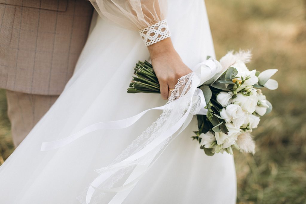 Close-up of a wedding couple. The focus is on the bride's hand holding a bouquet of white roses