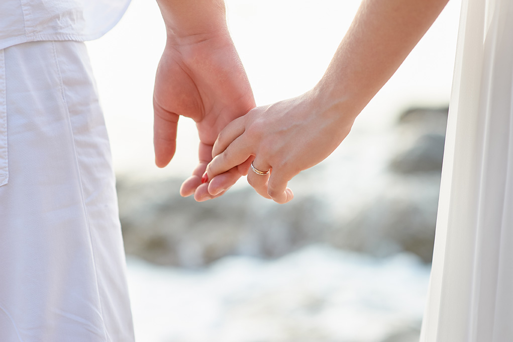 Two people dressed in white holding hands with a wedding ring as the center focus